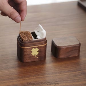 Pode incluir: Um porta-palitos de madeira com um design de trevo de quatro folhas. O suporte quadrado tem uma tampa e contém palitos e um pequeno pente branco. Uma mão segura um palito. O texto "MagicalDIYDesign" está na parte inferior.