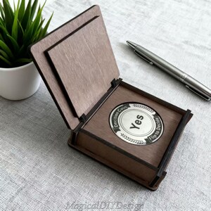 May include: A wooden decision coin box with the lid open, revealing a silver coin that says "Yes". The box is brown with a dark brown trim. A silver pen rests on the surface next to the box. A small green plant in a white pot is in the background.