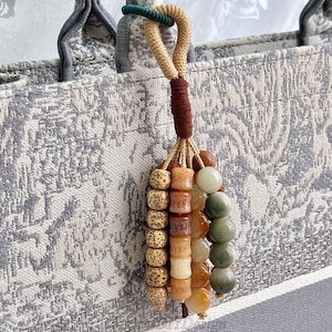 Könnte beinhalten: Ein dekorativer Taschenanhänger mit einer Vielzahl von Perlen in Braun-, Beige- und Grüntönen. Der Anhänger ist an einem beigen Seil und einem dunkelbraunen Akzent befestigt. Der Hintergrund ist ein grau-weiß gemusterter Stoff.