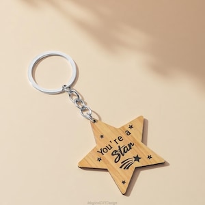 May include: Wooden star keyring with a silver ring and chain. The star features the text "You're a Star" in black, with star and shooting star graphics. The keyring is set against a light beige backdrop.