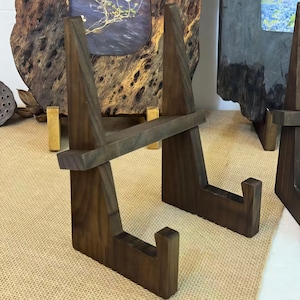 Puede incluir: Expositores de madera marrón oscuro. Los expositores tienen forma triangular con una pieza de soporte horizontal. Están diseñados para sostener y exhibir obras de arte u otros objetos. Fabricados en madera maciza.