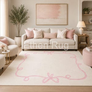 Puede incluir: Una alfombra color crema con un borde rosa de lazo y cinta. La alfombra está en un salón con un sofá, sillas y una pintura. La habitación tiene una paleta de colores neutros con acentos rosas.