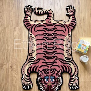 Puede incluir: Una alfombra rosa y negra con forma de tigre, con una cara detallada y un patrón de rayas. La alfombra está sobre un suelo de madera, con un pequeño libro y una taza blanca cerca. El diseño de la alfombra recuerda a una piel de tigre.
