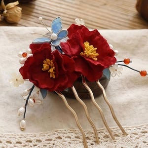 May include: A decorative hair comb featuring two large red flowers with yellow centers, a blue butterfly, and small white flowers. The comb has four gold-colored prongs, perfect for adding a touch of elegance to any hairstyle.
