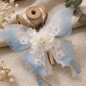 Könnte beinhalten: Zarte Haarspange in Schmetterlingsform mit durchscheinenden blau-weißen Flügeln. Die Mitte zeigt weiße Stoffblumen und kleine, weiße Blumenakzente. Ein goldfarbener Clip sichert das Accessoire.