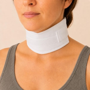 May include: A white cervical neck collar featuring a smooth fabric exterior and a horizontal hook-and-loop fastener strap. The medical support device wraps securely around the neck, providing a firm, structured fit with clean, finished edges.