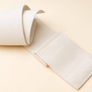 May include: A rolled-up, off-white elastic bandage with a ribbed texture. The bandage is partially unrolled, revealing a rectangular end with a smooth, padded section. The background is a light beige.