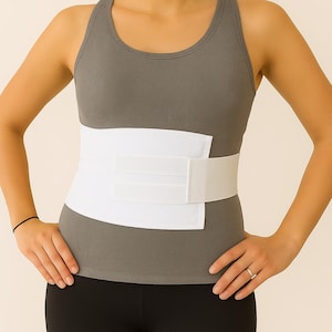May include: A white abdominal support band with a hook and loop closure is worn over a gray tank top. The band is designed to provide support and compression. The person is wearing black leggings.
