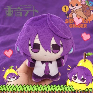 May include: A plush doll featuring purple hair, a white shirt with a purple tie, and purple accents. The doll has a neutral expression with dark eyes. The background includes graphics of a fox, a yellow pear, and pixelated hearts and grass.
