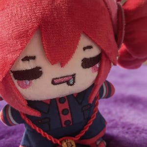 May include: A plush doll with bright red hair, featuring embroidered closed eyes, a small mouth with a tongue sticking out, and pink cheeks. The doll wears a dark blue outfit with red trim, buttons, and a red cord necklace with a small wooden bead.
