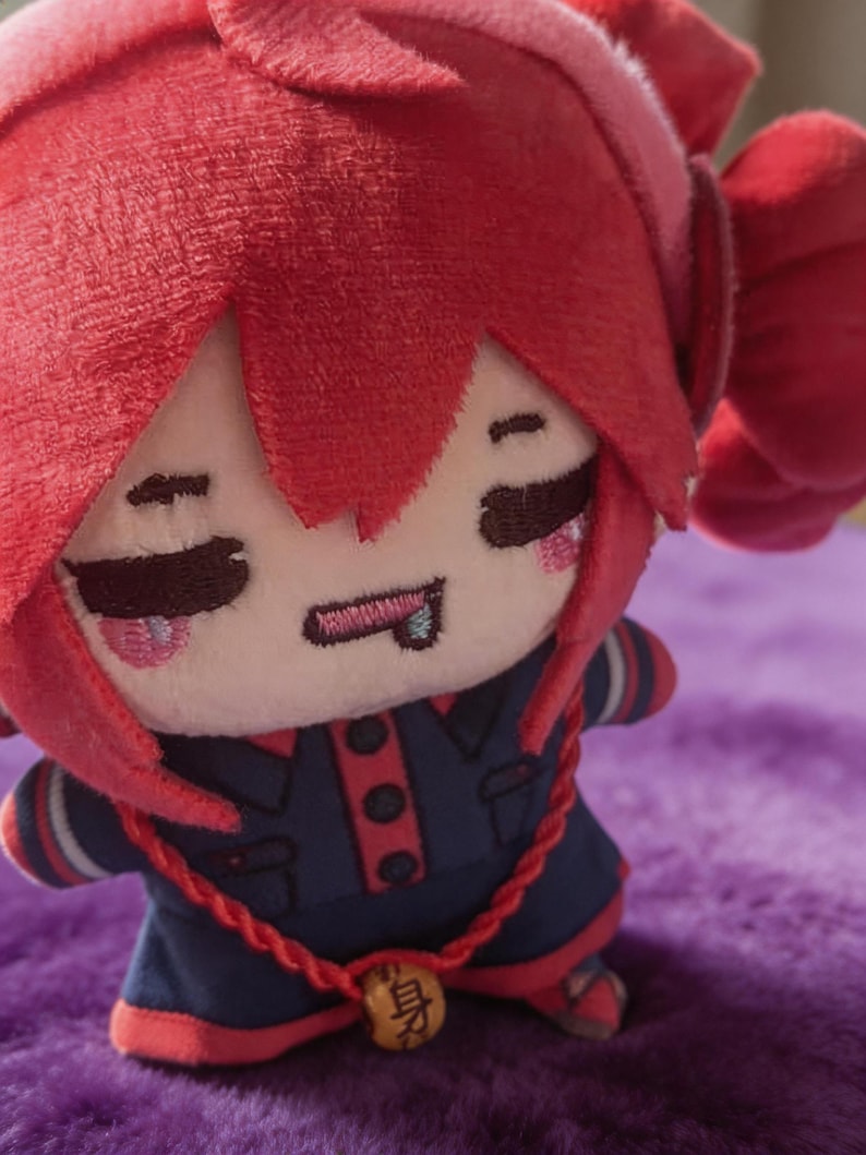 May include: A plush doll with bright red hair, featuring embroidered closed eyes, a small mouth with a tongue sticking out, and pink cheeks. The doll wears a dark blue outfit with red trim, buttons, and a red cord necklace with a small wooden bead.