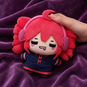 May include: A plush toy featuring a character with bright pink hair styled in twin tails, wearing pink earmuffs and a dark blue collared outfit with red accents. The character has closed eyes, pink blush marks, and a small blue drool detail on its mouth.
