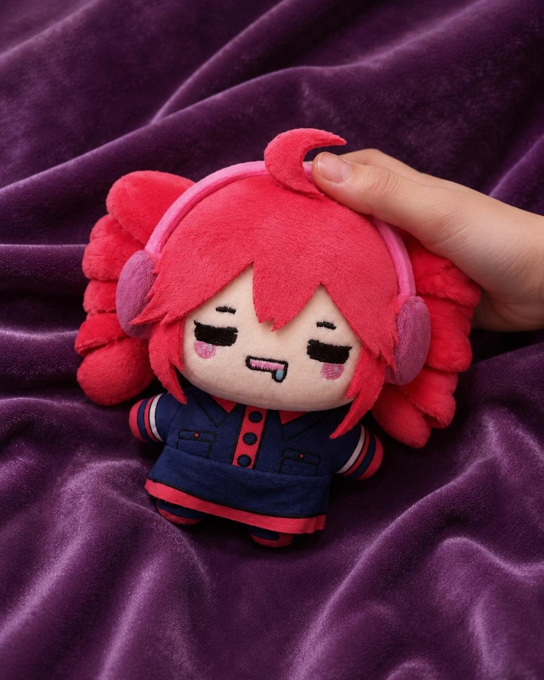 May include: A plush toy featuring a character with bright pink hair styled in twin tails, wearing pink earmuffs and a dark blue collared outfit with red accents. The character has closed eyes, pink blush marks, and a small blue drool detail on its mouth.