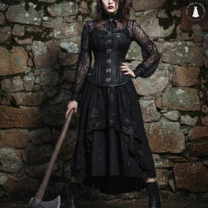 Może przedstawiać: Czarny gotycki strój składający się z koronkowej bluzki, gorsetu i warstwowej spódnicy, w połączeniu z czarnymi butami i toporem. Widoczne logo The Dark Wardrobe Co. Scena rozgrywa się na tle kamiennej ściany.