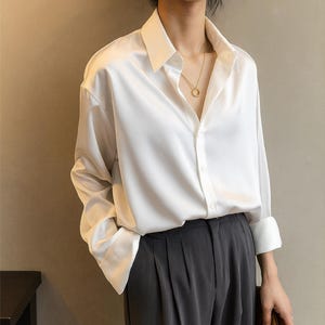 Puede incluir: Una camisa blanca de satén con botones, mangas largas y cuello clásico, combinada con pantalones plisados grises oscuros. Un collar dorado y un bolso de mano marrón completan el look. Adecuado para negocios o uso casual.