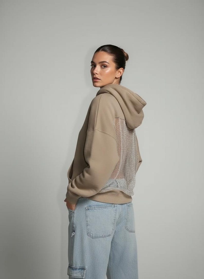 K&ouml;nnte beinhalten: Ein beigefarbener Kapuzenpullover mit Mesh-R&uuml;ckendetail. Der Hoodie hat eine Kapuze mit Kordelzug und eine K&auml;ngurutasche. Das Model tr&auml;gt eine Jeans in heller Waschung. Der Stil ist l&auml;ssig und modern.