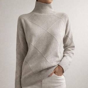 Op de afbeelding: Een lichtbeige coltrui met een ruitpatroon. De trui heeft een geribbelde coltrui en een ruitontwerp op de voorkant en mouwen. Het model draagt een witte broek en heeft de hand in de zak.