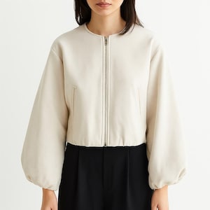May include: Cream-colored jacket with a round neckline and a silver zipper. The jacket has long, voluminous sleeves and two small pockets. The model is wearing black pants. The jacket is a neutral color and could be worn with many outfits.
