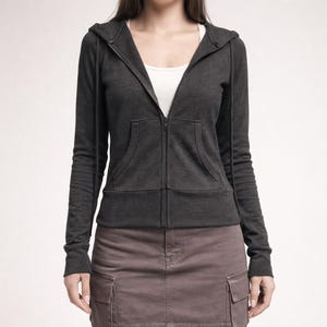 May include: A dark grey zip-up hoodie with a drawstring hood and front pockets. The hoodie is worn over a white vest top. The model is also wearing a mauve cargo skirt.