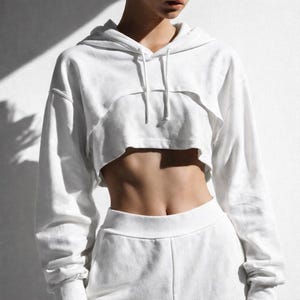 May include: White cropped hoodie with a drawstring hood and long sleeves. The hoodie is paired with matching white sweatpants. The outfit is made of a soft, comfortable material. The cropped design reveals the midriff.