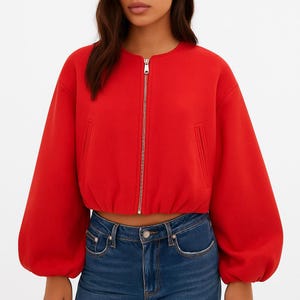 May include: A bright red bomber jacket with a silver zipper and two side pockets. The jacket has a round neckline and long, voluminous sleeves. The bottom hem is elasticized. The model is wearing blue jeans.