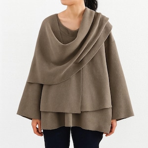 May include: A taupe-colored cape-style top with a draped cowl neckline and layered design. The top has wide sleeves and a relaxed fit. The model is wearing dark blue pants. The fabric appears to be a soft, woven material.