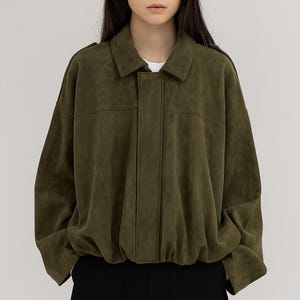 May include: Olive green jacket with a collared neckline and a button-up front. The jacket has a relaxed fit with long sleeves and a gathered hem. The model is wearing a white shirt underneath and black pants.