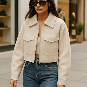 May include: A woman wearing a beige cropped jacket with two front pockets, a matching crop top, and blue jeans. The jacket has a collar and a zipper closure. She is also wearing sunglasses.
