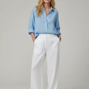 May include: A woman wearing a light blue button-down shirt and white wide-leg pants. The shirt has long sleeves and a collared neckline. The pants are high-waisted and have pockets. The woman is also wearing sunglasses.
