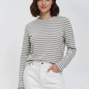 Pode incluir: Uma camisa de manga comprida com riscas horizontais cinzentas e brancas. A camisa é combinada com jeans brancos. A camisa tem gola redonda e um padrão de riscas clássico. A modelo está a usar a camisa e os jeans.
