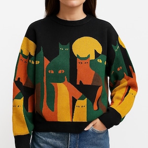 May include: Black crew neck jumper with a colourful cat design. The design features orange, green, and yellow cats with a yellow moon against a black background. The jumper has black cuffs and a black hem.
