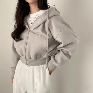 Op de afbeelding: Een lichtgrijze cropped hoodie met een capuchon met trekkoord en een ritssluiting aan de voorkant. De hoodie heeft een kangoeroezak en geribde manchetten en zoom. Het model draagt een wit T-shirt en een witte broek.