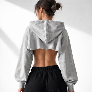 May include: A gray cropped hoodie with long sleeves and a raw hem. The back of the hoodie is open, revealing the wearer's back. The sleeves are long and loose, and the hoodie has a hood. The person is wearing black sweatpants.