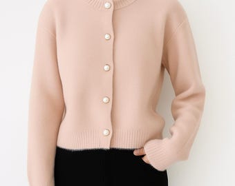 May include: A light pink cardigan with a crew neck and long sleeves. The sweater has five pearl-like buttons down the front. The bottom half of the person is wearing black pants.