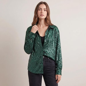 Könnte beinhalten: Ein langärmliges, smaragdgrünes Paillettenhemd mit Kragen. Das Hemd ist zugeknöpft und wird mit einer schwarzen Jeans kombiniert. Das Model trägt ein schwarzes Top unter dem Hemd.