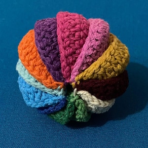 May include: A colorful, handmade crochet ball featuring a variety of vibrant colors, including orange, purple, pink, and green. The ball is made of yarn and has a textured surface. The crochet ball is a decorative item.