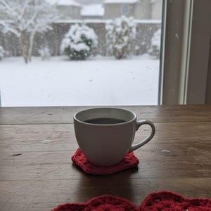 May include: A white ceramic mug filled with dark liquid sits on a red crocheted heart-shaped coaster. Two additional red heart coasters are on a wooden table. A snowy outdoor scene is visible through a window.