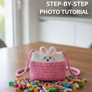 May include: A handmade crochet bunny bag in white and pink, with a bunny face and ears. The bag has a pink strap and is surrounded by colorful candies. The image includes the text "STEP-BY-STEP PHOTO TUTORIAL".