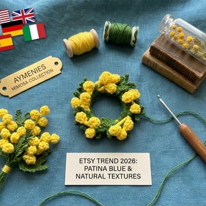 Op de afbeelding: Een verzameling handgemaakte items met mimosa-thema, waaronder een gehaakte krans en boeket, klosjes garen en een kleine glazen fles met mimosatakken. Een haaknaald en een label met "AYMENIES MIMOSA COLLECTION" zijn ook zichtbaar.