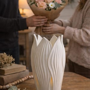 May include: A white ceramic vase with a tulip-like design, featuring vertical, curved lines. The vase is on a wooden table, with a bouquet of pink and white flowers being handed to someone in the background. Decorative home decor.