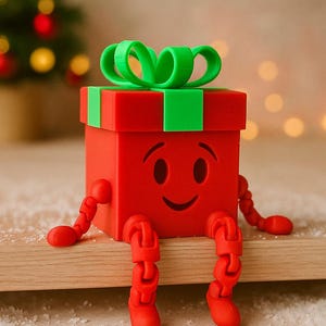 May include: A red gift box with a green bow and accents, featuring a smiling face and articulated legs. The festive decoration is sitting on a wooden surface, with a Christmas tree in the background.