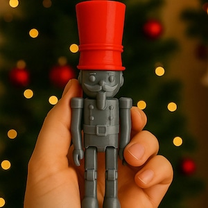 Puede incluir: Una figura de soldado de juguete gris con un sombrero rojo. La figura tiene bigote y se sostiene en una mano. El fondo es un árbol de Navidad borroso con adornos rojos y luces amarillas.