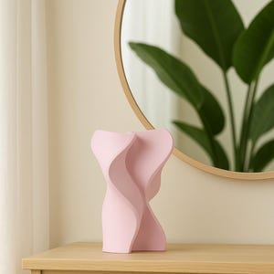 May include: A light pink, abstract ceramic vase with a wavy design. The vase is displayed on a light wood surface, with a round mirror and green plant in the background. The vase is approximately 10 inches tall.