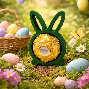 May include: A green bunny-shaped holder cradles a Ferrero Rocher chocolate. The scene is set outdoors with a basket of Easter eggs, painted eggs, flowers, and a butterfly. Pastel-colored eggs hang from the trees.