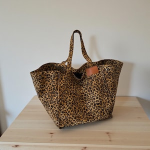 May include: A large tote bag with a leopard print pattern. The bag has two handles and a small brown leather tag. The bag is sitting on a light brown wooden surface.