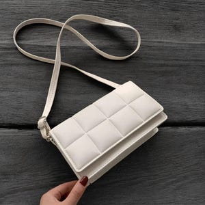 以下が含まれることがあります： 白のキルティング加工されたクロスボディバッグ。細いストラップが付いており、長さを調節できます。バッグは長方形で、フラップ開閉式です。バッグは暗い木製の表面に置かれています。