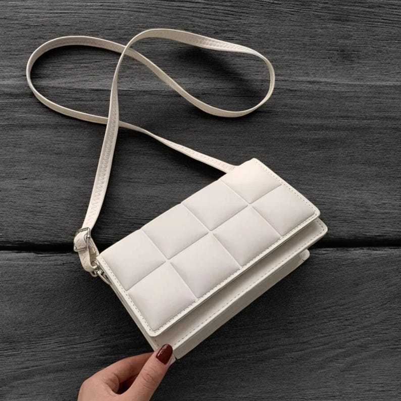 以下が含まれることがあります： 白のキルティング加工されたクロスボディバッグ。細いストラップが付いており、長さを調節できます。バッグは長方形で、フラップ開閉式です。バッグは暗い木製の表面に置かれています。