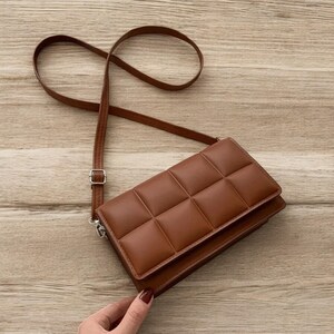 以下が含まれることがあります： 長い調節可能なストラップが付いた、茶色のキルティングクロスボディバッグ。 バッグは長方形で、フラップ開閉とシルバーカラーの金具が付いています。 バッグは滑らかな革のような素材で作られています。