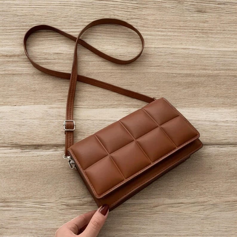 以下が含まれることがあります： 長い調節可能なストラップが付いた、茶色のキルティングクロスボディバッグ。 バッグは長方形で、フラップ開閉とシルバーカラーの金具が付いています。 バッグは滑らかな革のような素材で作られています。
