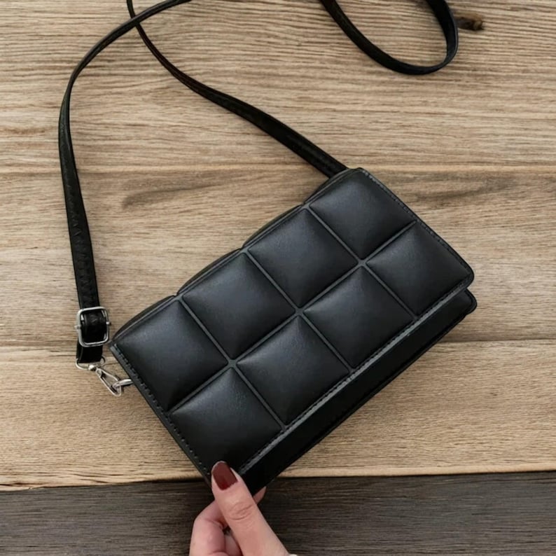 以下が含まれることがあります： キルティングデザインの黒いクロスボディバッグ。長くて調節可能なストラップとシルバーカラーのバックルが付いています。長方形のバッグはフラップ開閉式です。背景は木製の表面です。